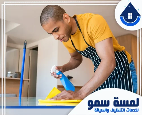 تنظيف قصور بالرياض عمالة فلبينية 0554516792 _ لمسة سمو يظهر موظف من فريق لمسة سمو وهو يقوم بمسح طاولة طعام أو سطح عمل بتركيز شديد. يرتدي العامل مئزراً مخططاً وقفازات زرقاء.